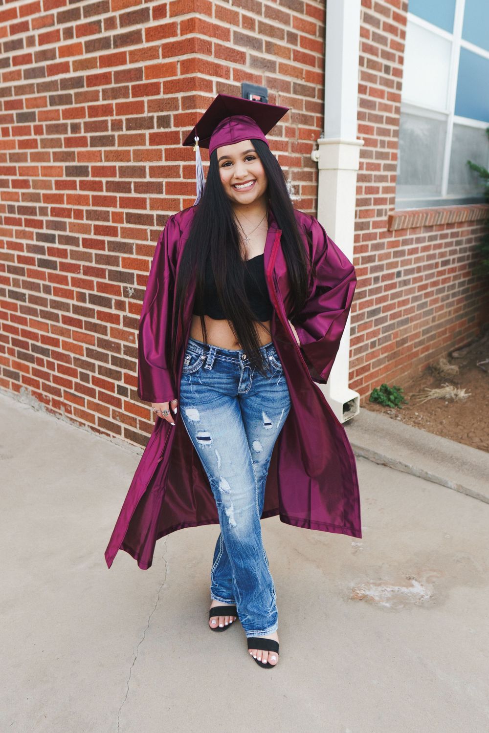Graduate in maroon cap and gown poses with ripped jeans and crop top against brick wall.
