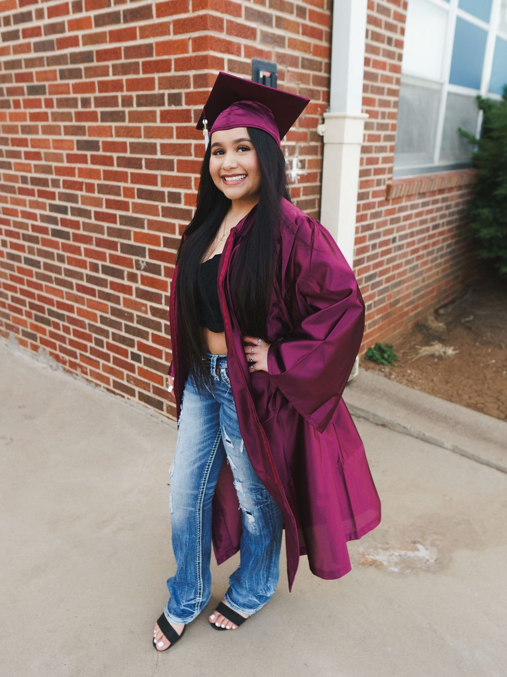Graduate wearing maroon graduation gown and cap poses confidently on sidewalk.
