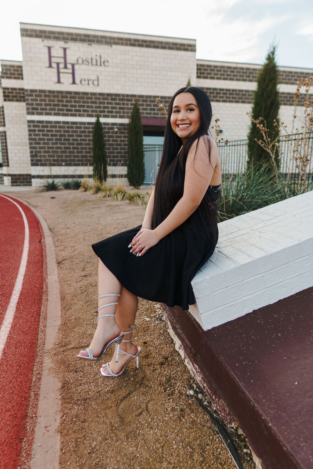 Woman in black dress poses near running track at sunset.
