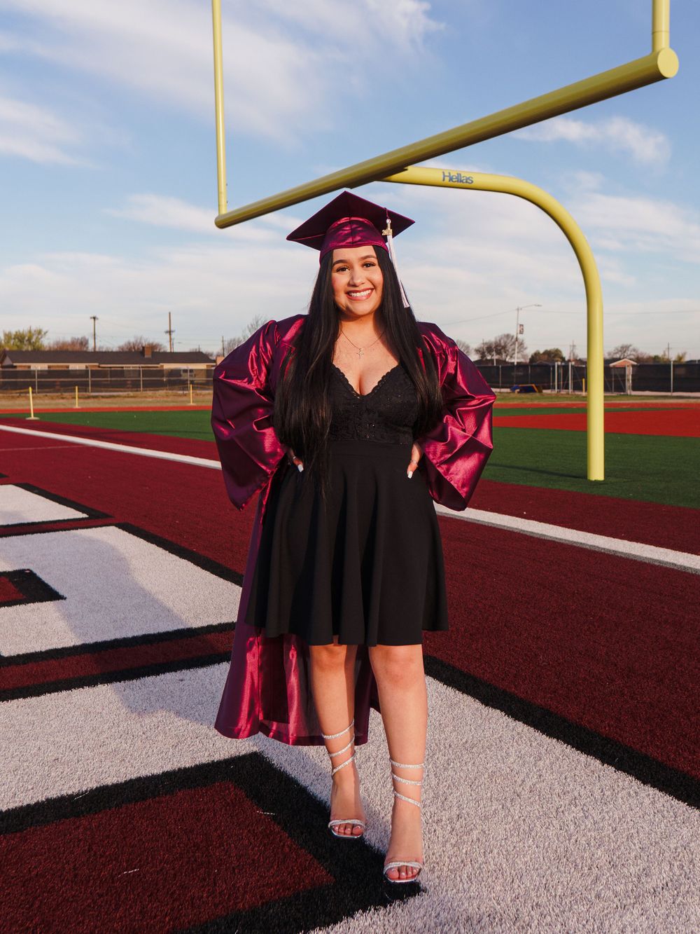 Graduate in maroon cap and gown poses under football goal post.