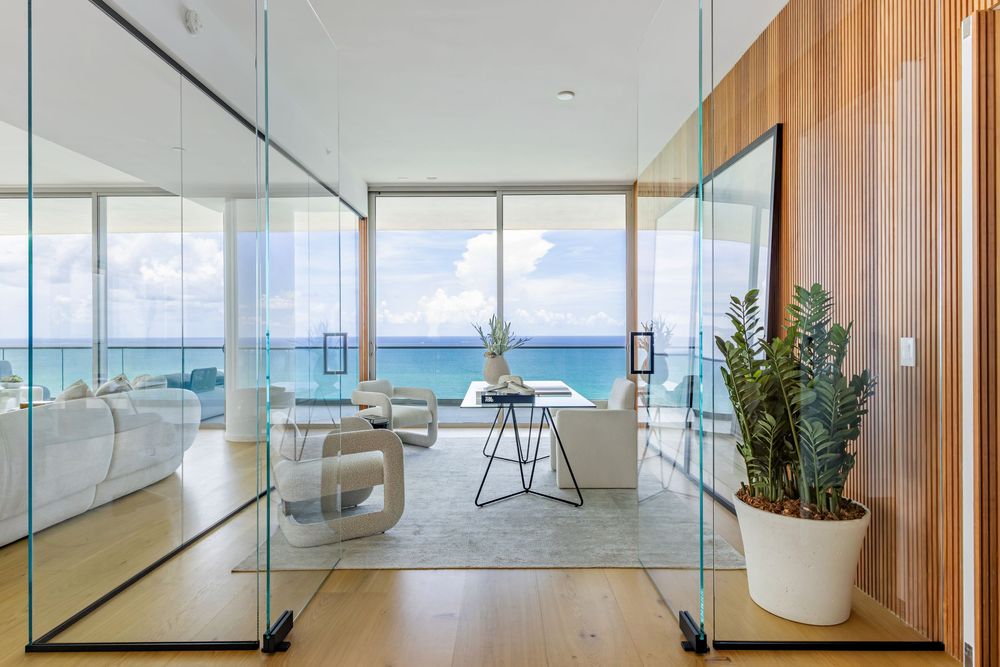 Luxurious beachfront living room with floor-to-ceiling windows offering panoramic ocean views and modern white furnishings.