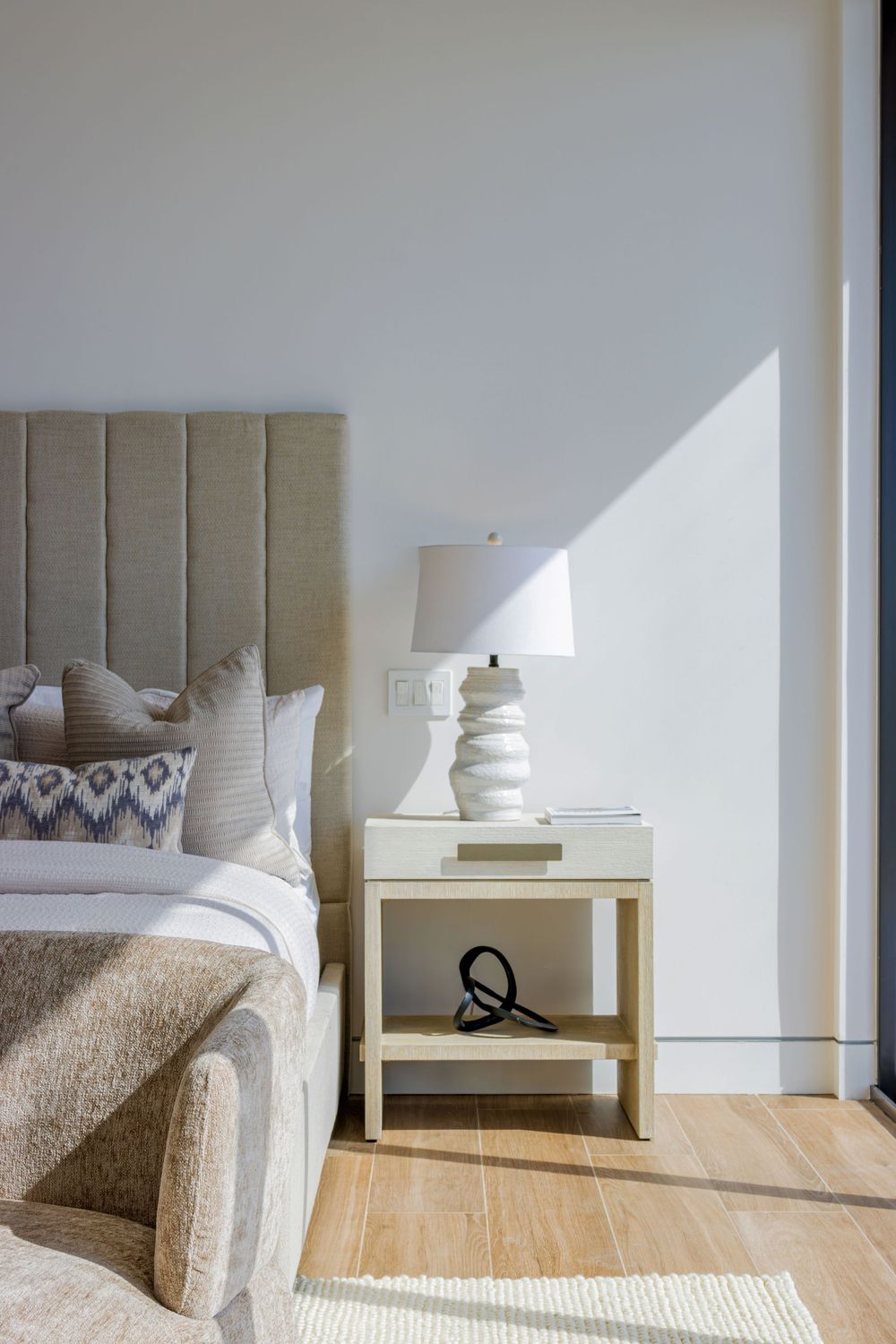 Minimalist bedroom corner with beige upholstered headboard and white bedside table in natural lighting.