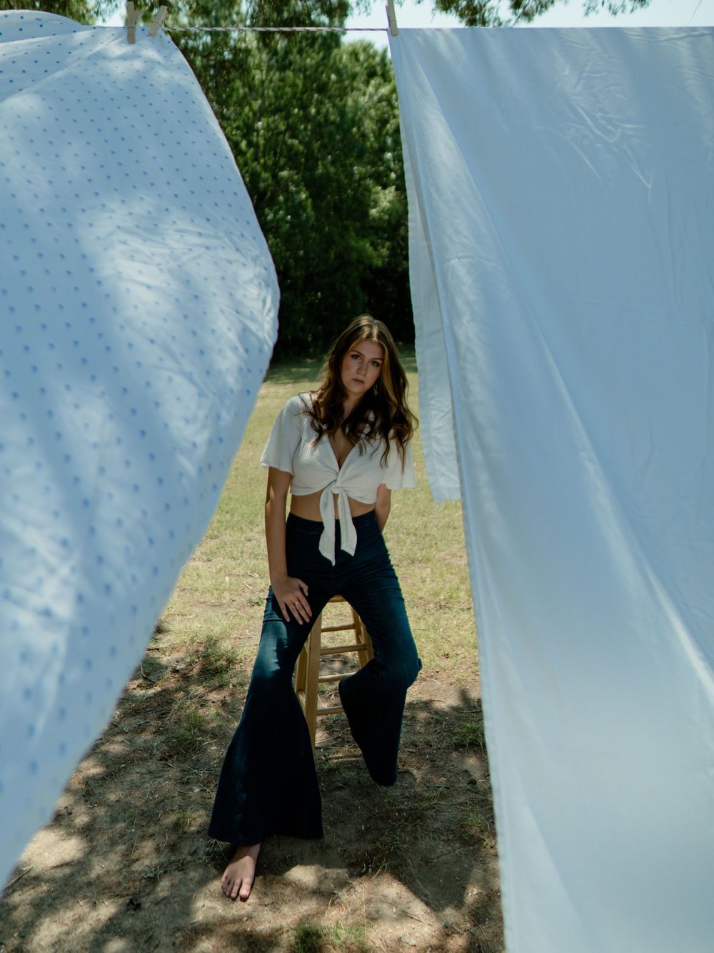 Model in white tied top and dark pants poses between white fabric panels outdoors.