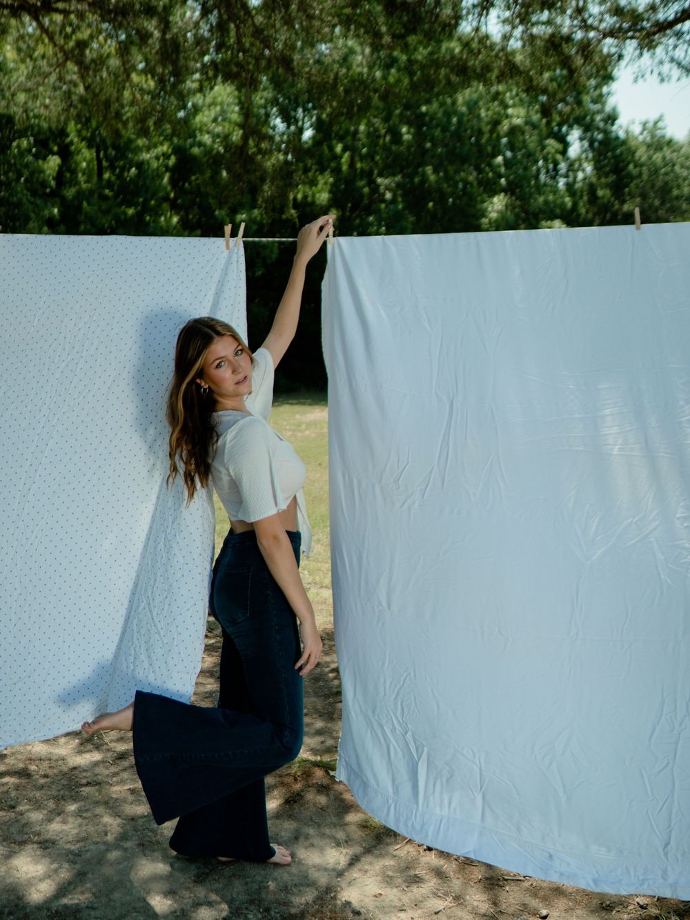 Model in white crop top and flare jeans poses playfully with white sheets hanging on clothesline outdoors.