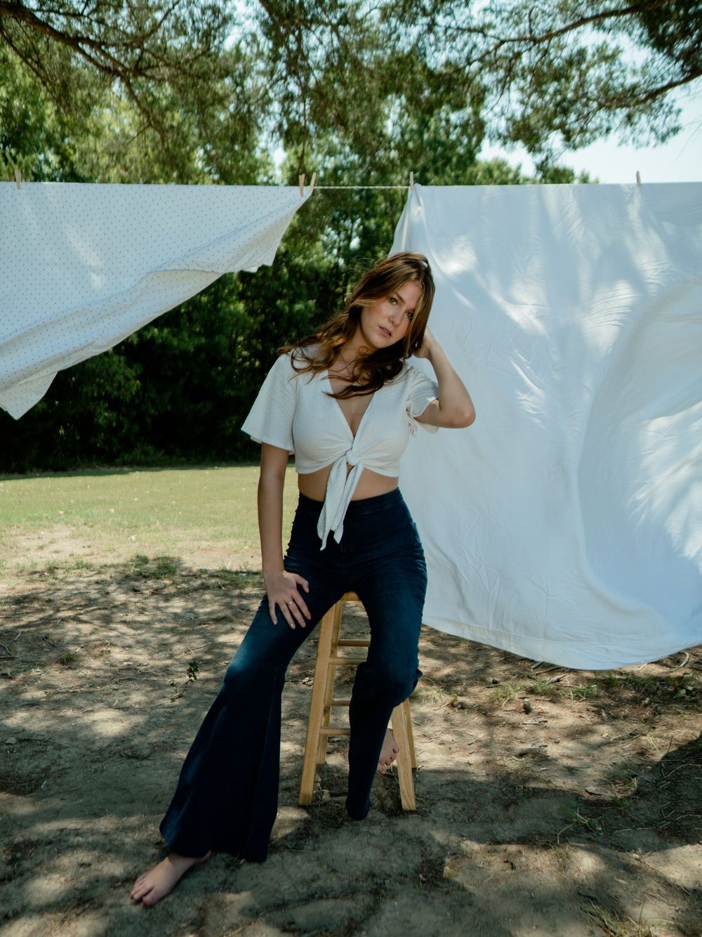 Fashion shoot with white tie-front top and navy flared pants against hanging white sheets in sunlit garden.