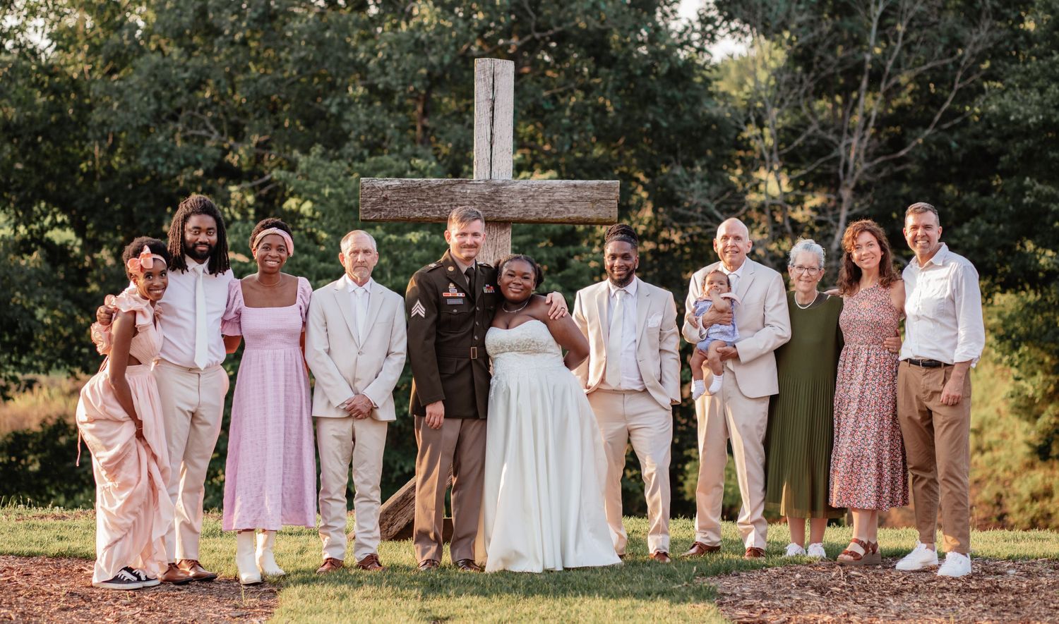 A large wedding party poses for a group photo in a garden setting wearing pastel colors.