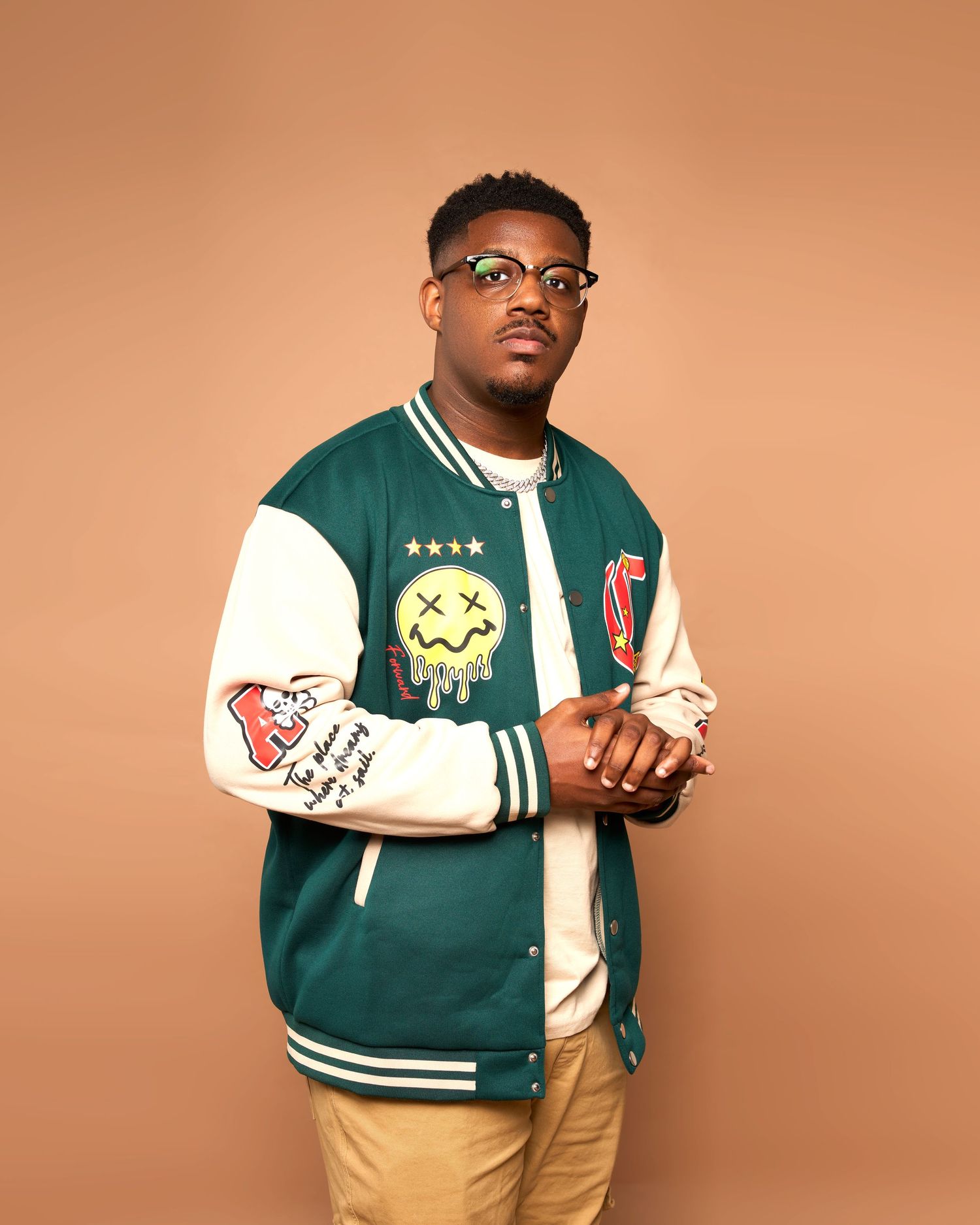 Portrait of person in green varsity jacket with patches and khaki pants against orange background.