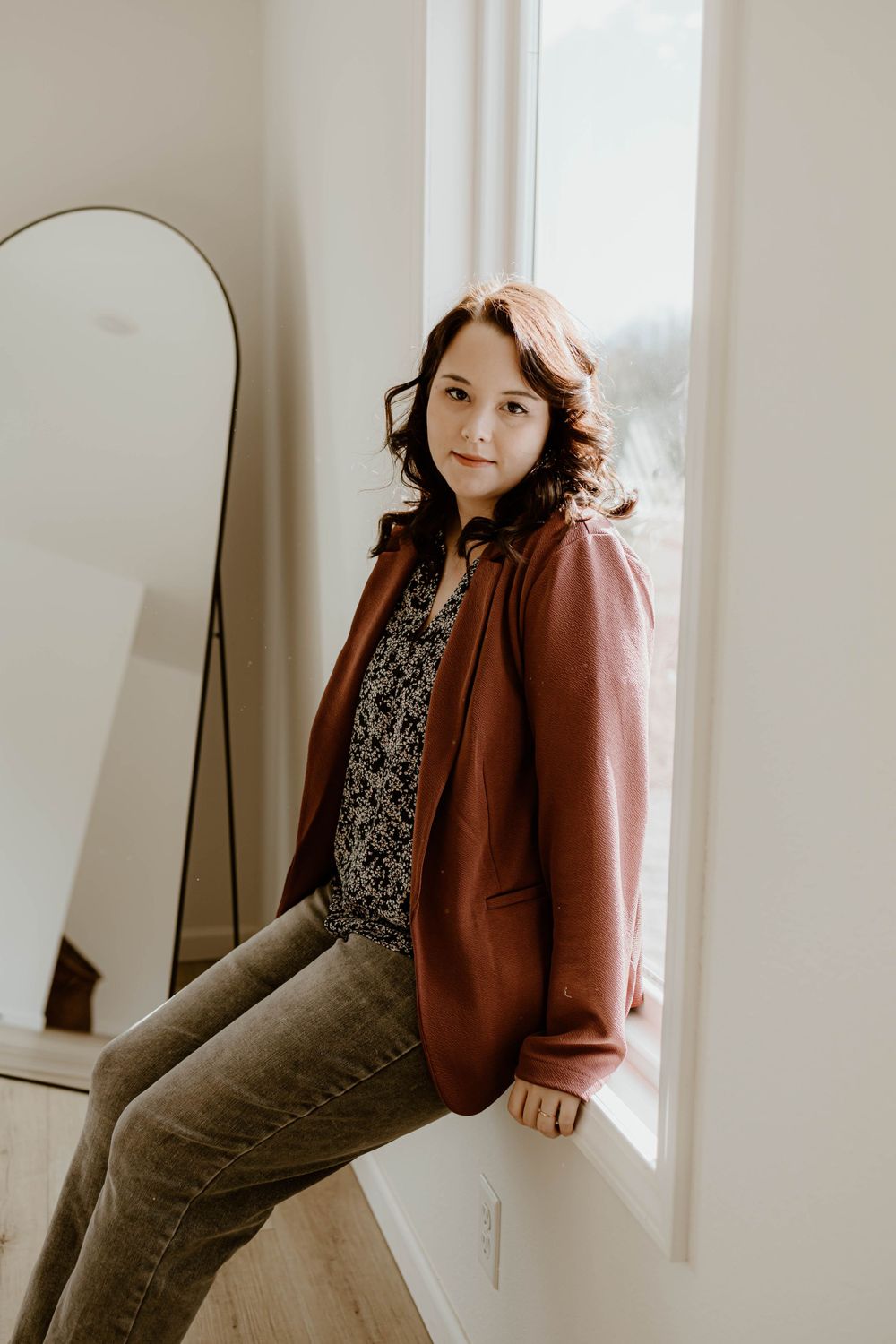 Professional fashion portrait of a person in a brown blazer and floral top posing against a white wall with mirror.