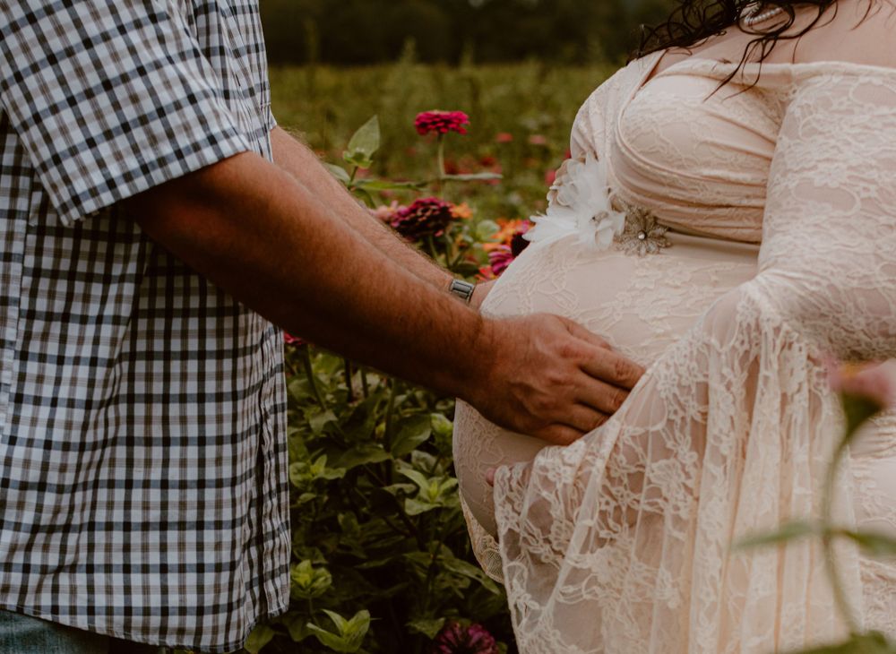 Close-up of hands touching pregnant belly in white lace dress among garden flowers.