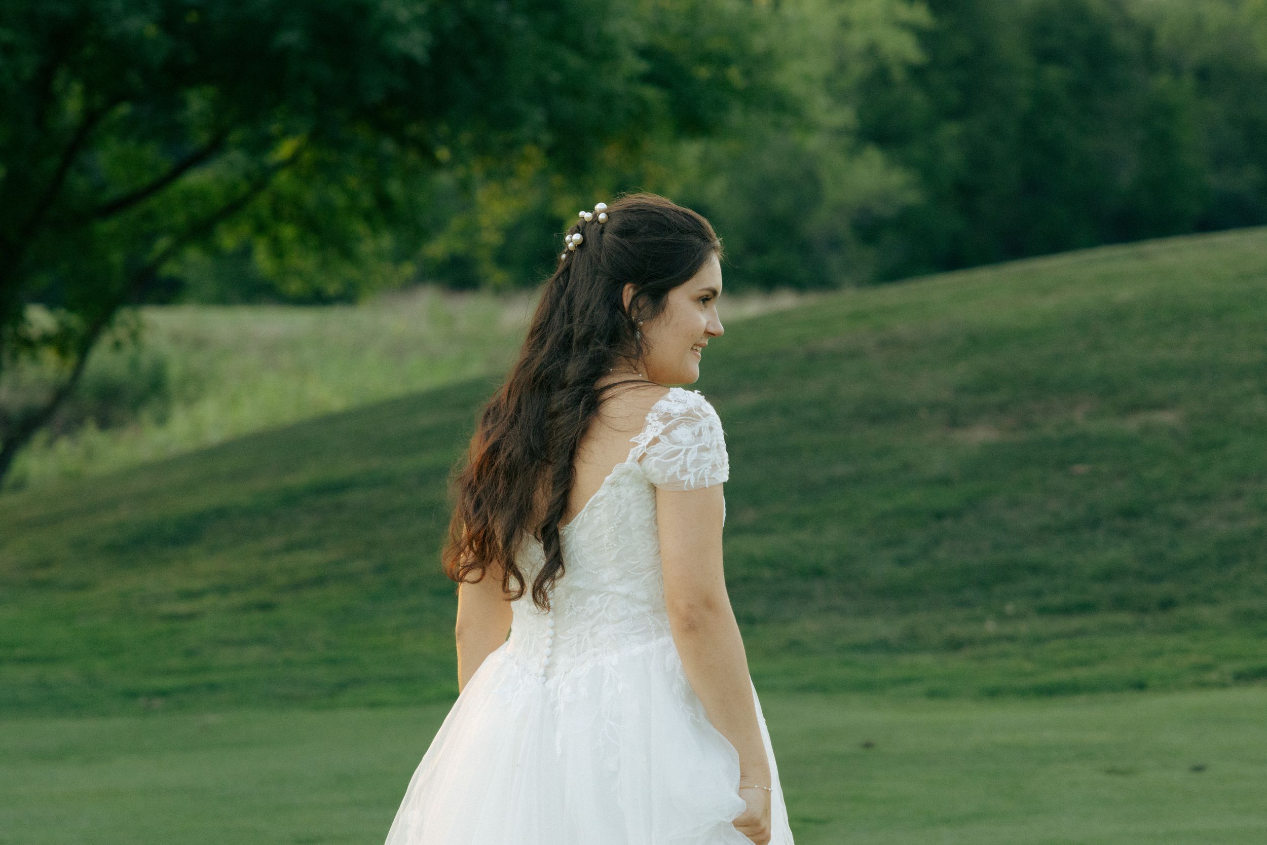 Bride in white lace dress with long dark hair stands on a grassy hillside during golden hour.