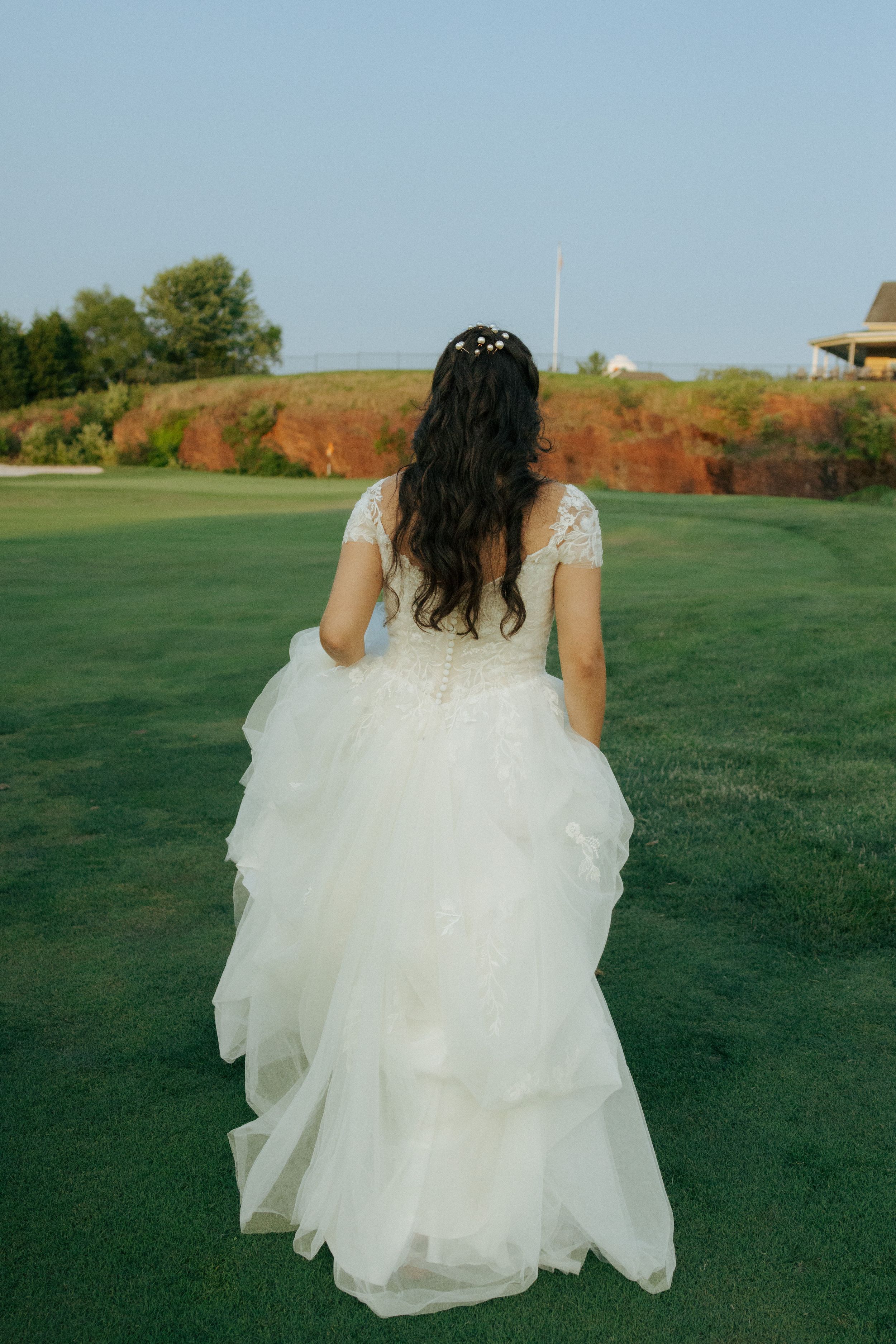 Back view of bride in flowing white gown walking across green lawn at sunset.