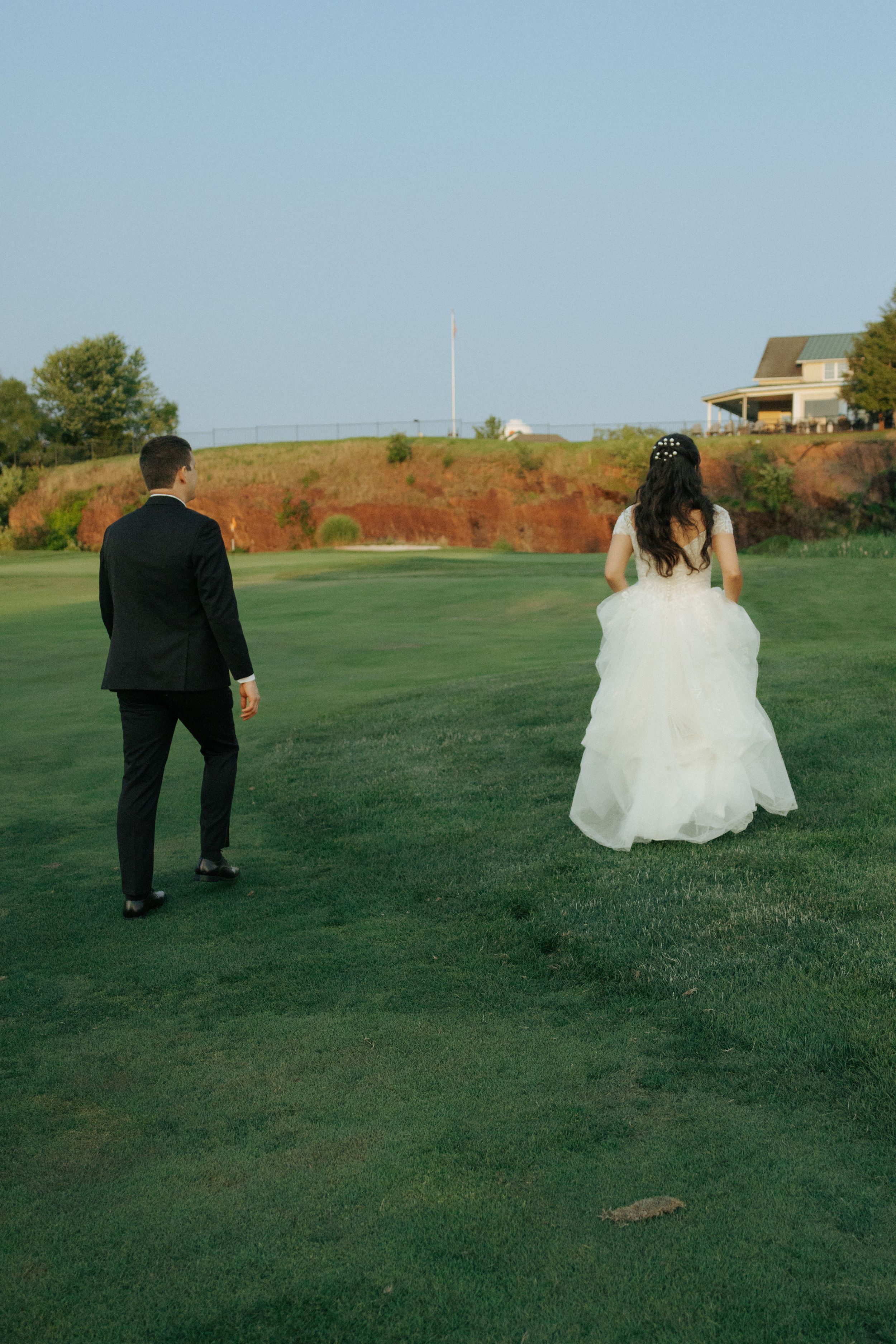 Bride and groom walking apart on a green lawn with houses on a hillside in the background.