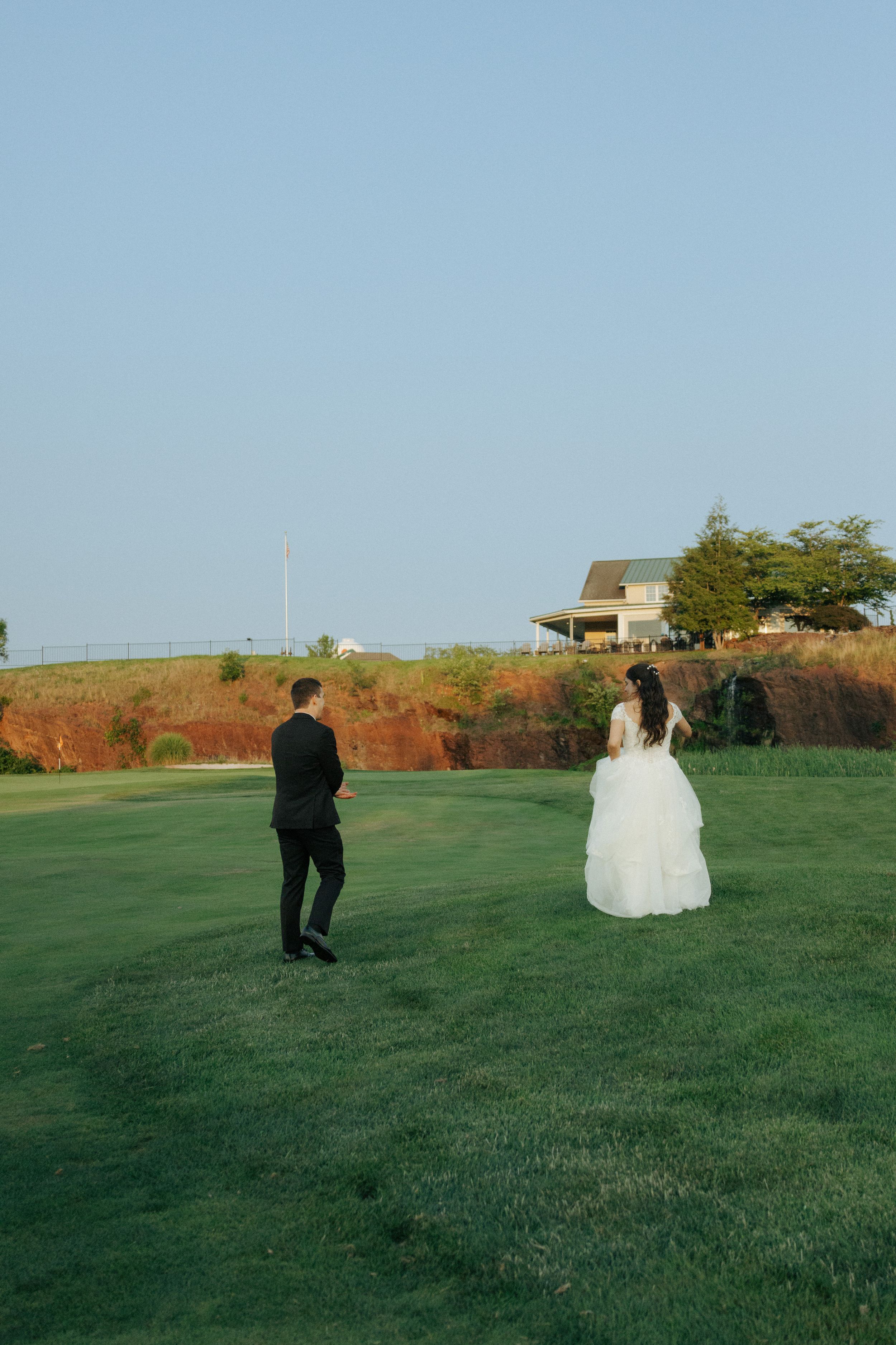 A couple in wedding attire walk across a grassy hilltop with a house in the background.