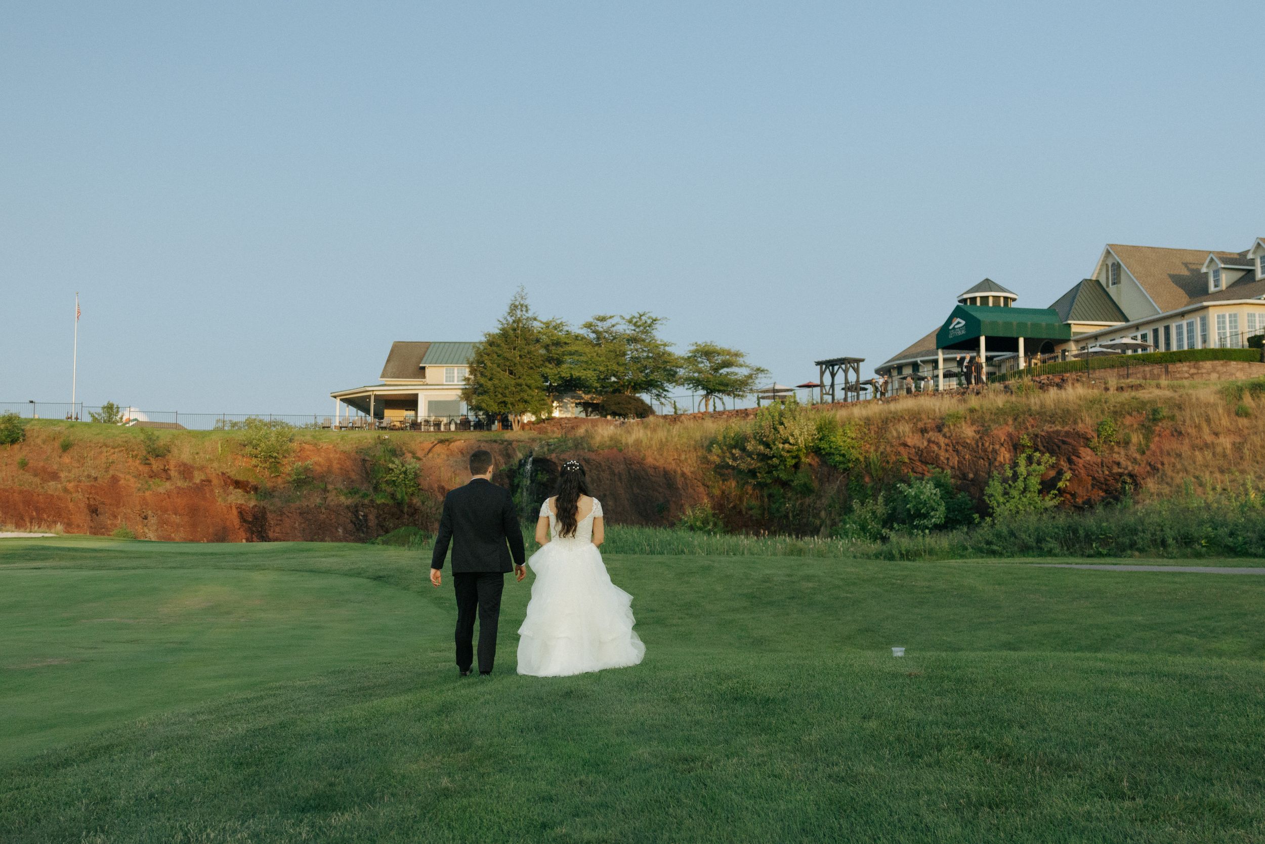 Bride and groom walking across a golf course with coastal homes on the hillside in background.