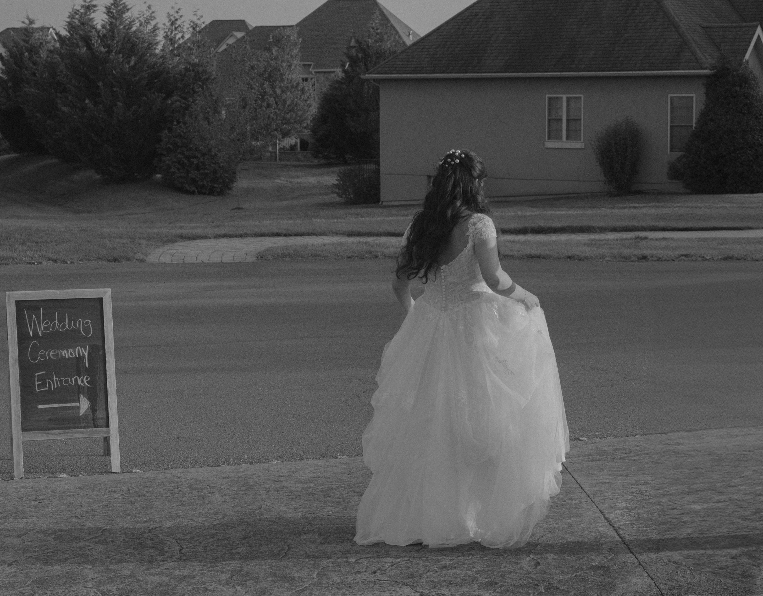 Black and white image of bride in full wedding gown walking past a sidewalk chalkboard sign.