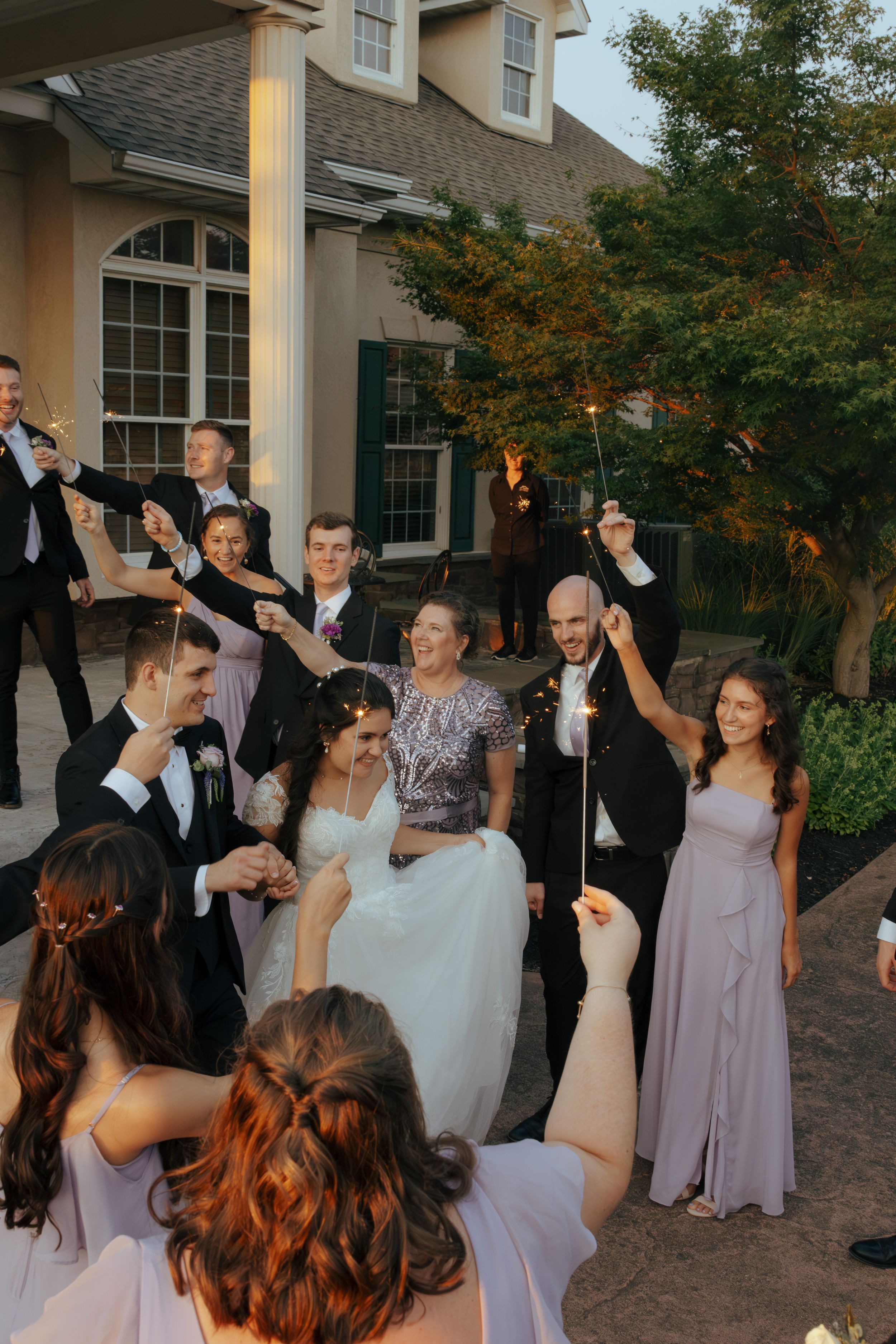Wedding party celebrates outdoors with raised arms in front of a house at dusk.