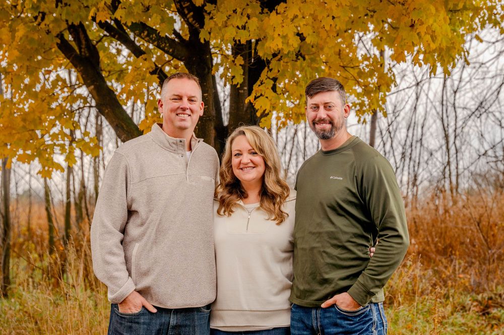 Three people in casual sweaters pose together under a golden autumn tree with fallen leaves in the background.
