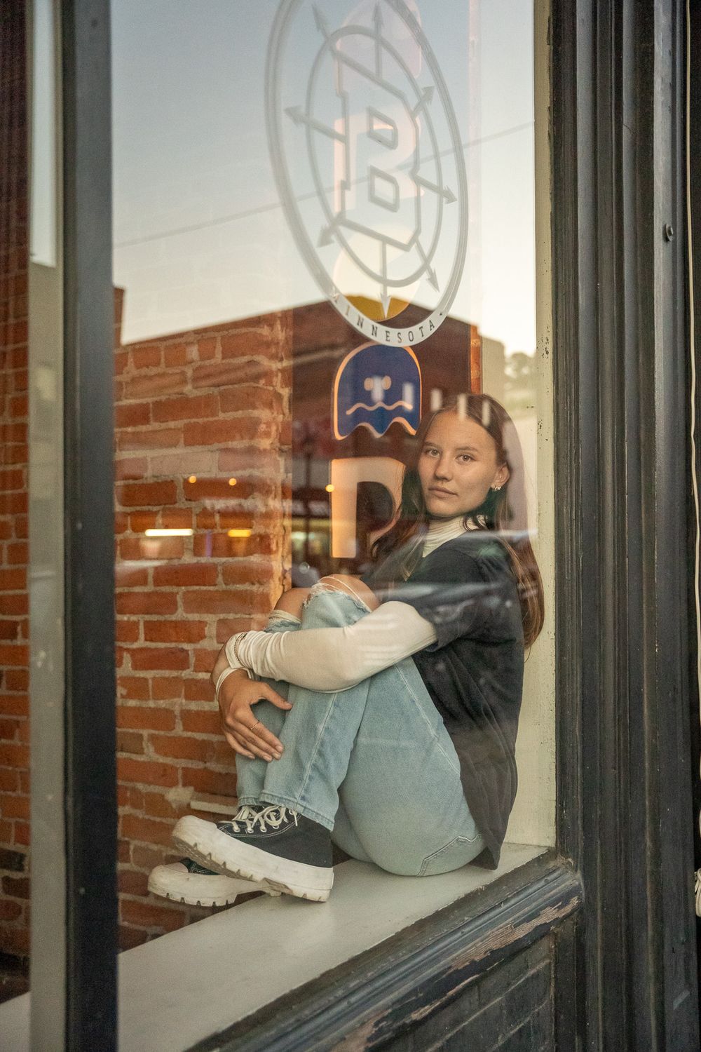 Someone sits in a window sill of a brick building with a neon sign visible in the background.