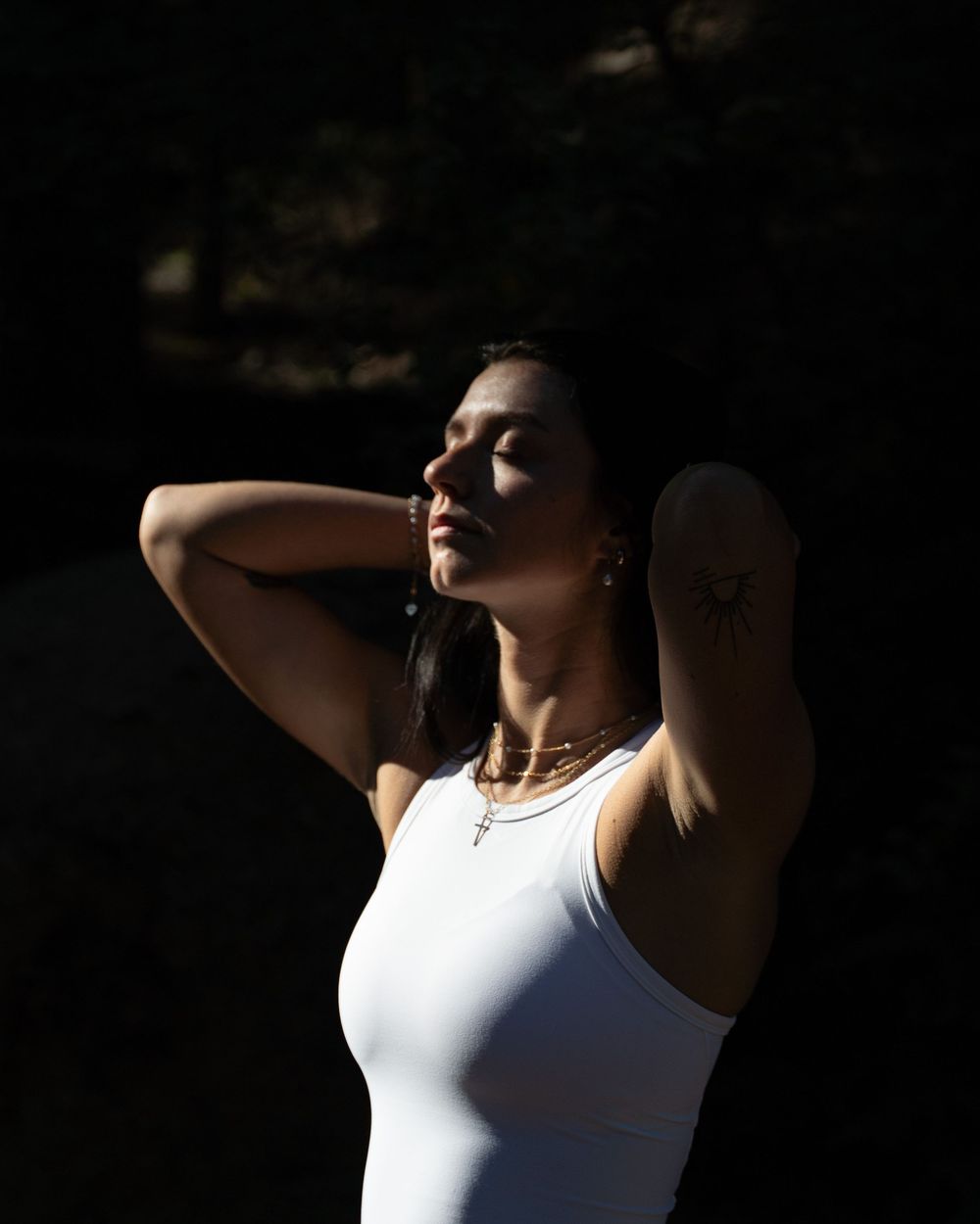 A person in a white tank top poses against a dark background with dramatic side lighting creating a moody silhouette effect.