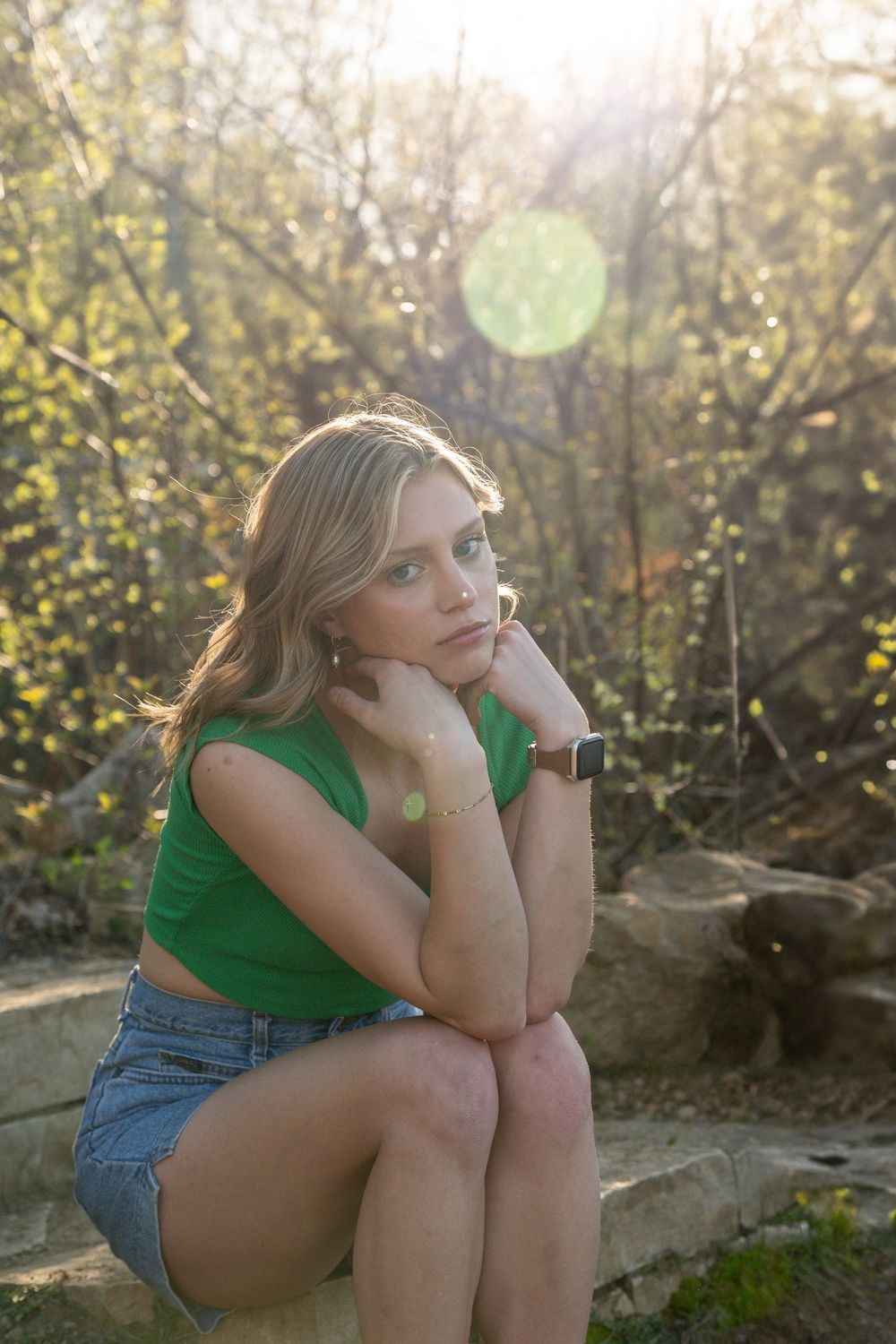 A person in a green top and denim sits thoughtfully on rocks during golden hour in a sunlit natural setting.