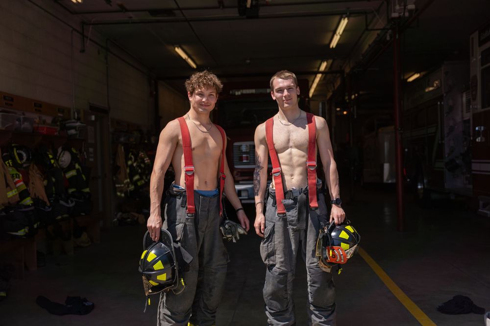 Two shirtless firefighters in suspenders and work pants stand side by side holding helmets in a dark station.