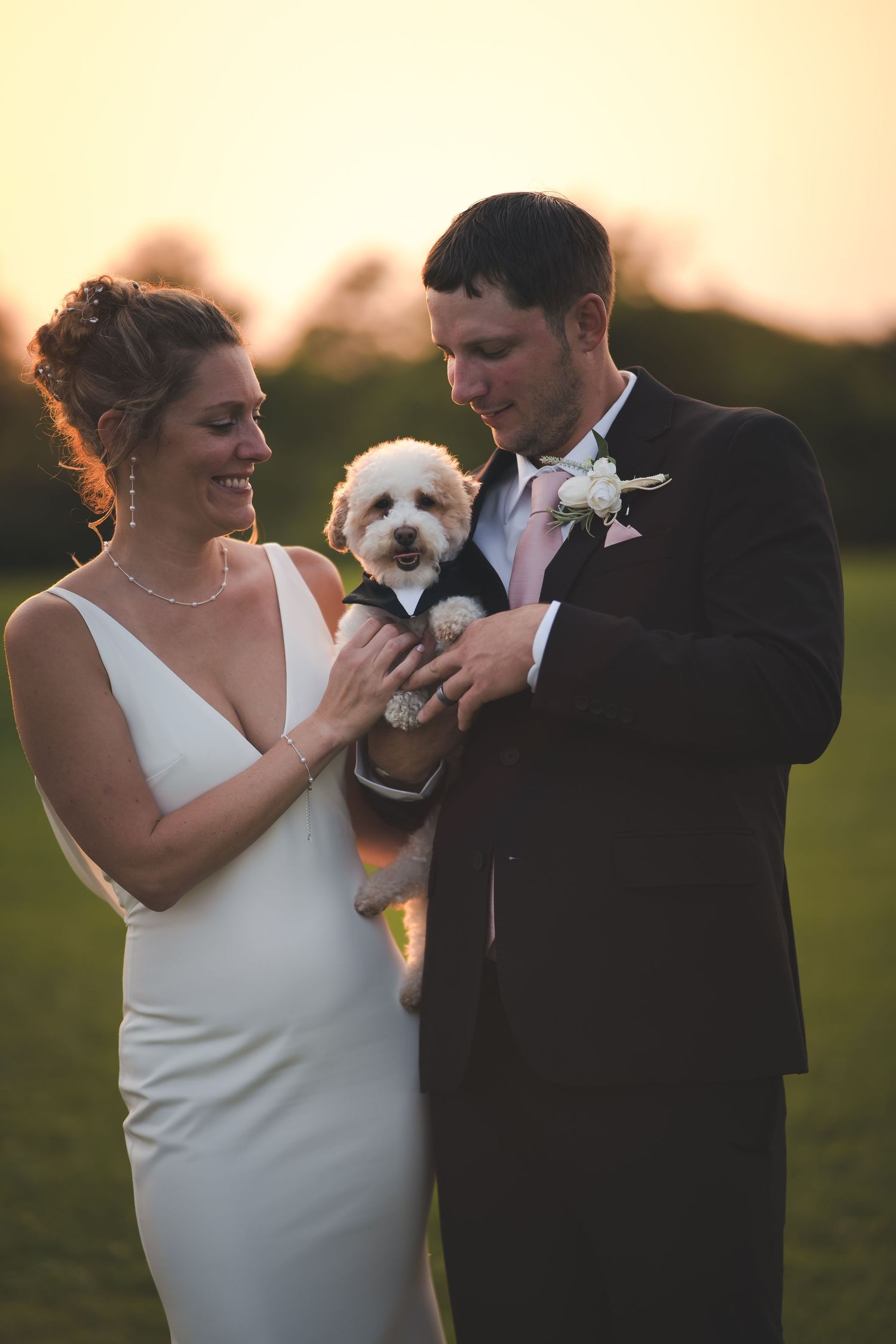 Newlyweds in formal attire holding their small white dog during sunset wedding photos.