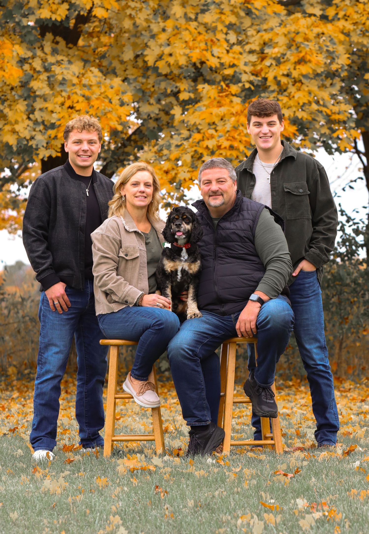 Casual family portrait with small black dog sitting on wooden stools against fall foliage.