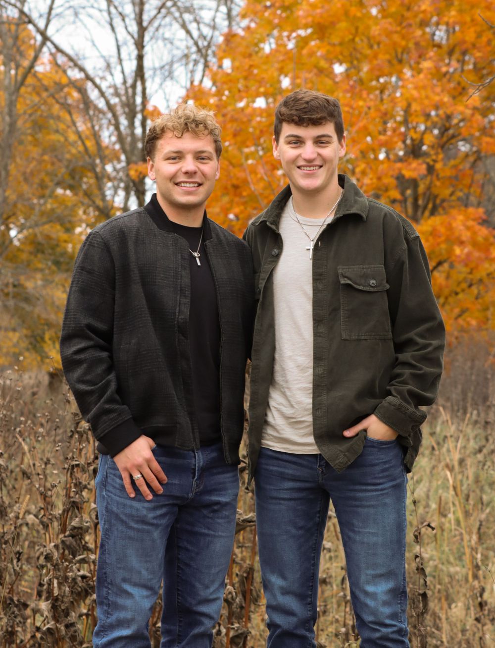 Two people in casual jackets pose together against colorful fall foliage backdrop.