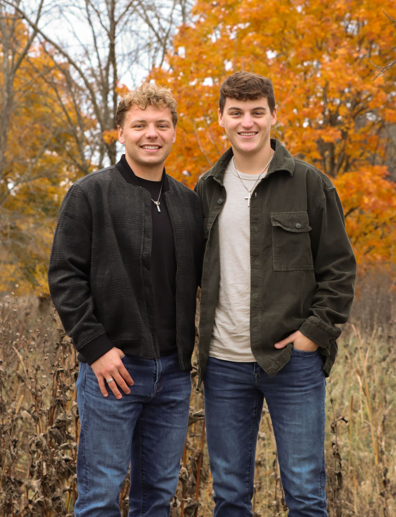 Two people in casual jackets pose together against colorful fall foliage backdrop.
