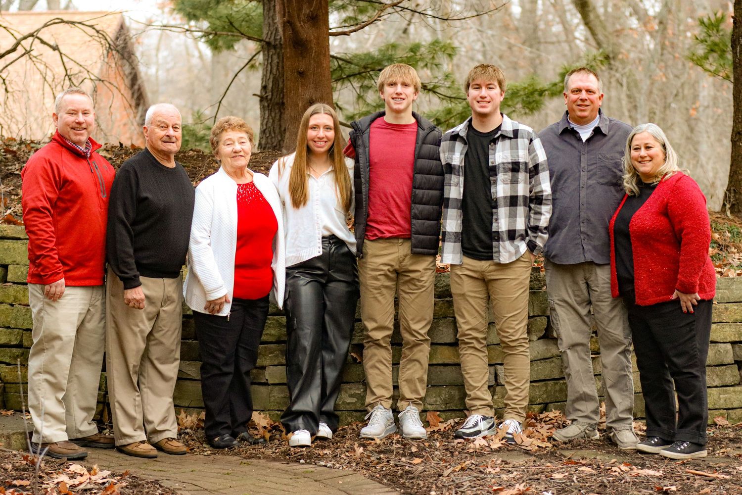 Family group portrait with eight people standing together against stone wall and trees.
