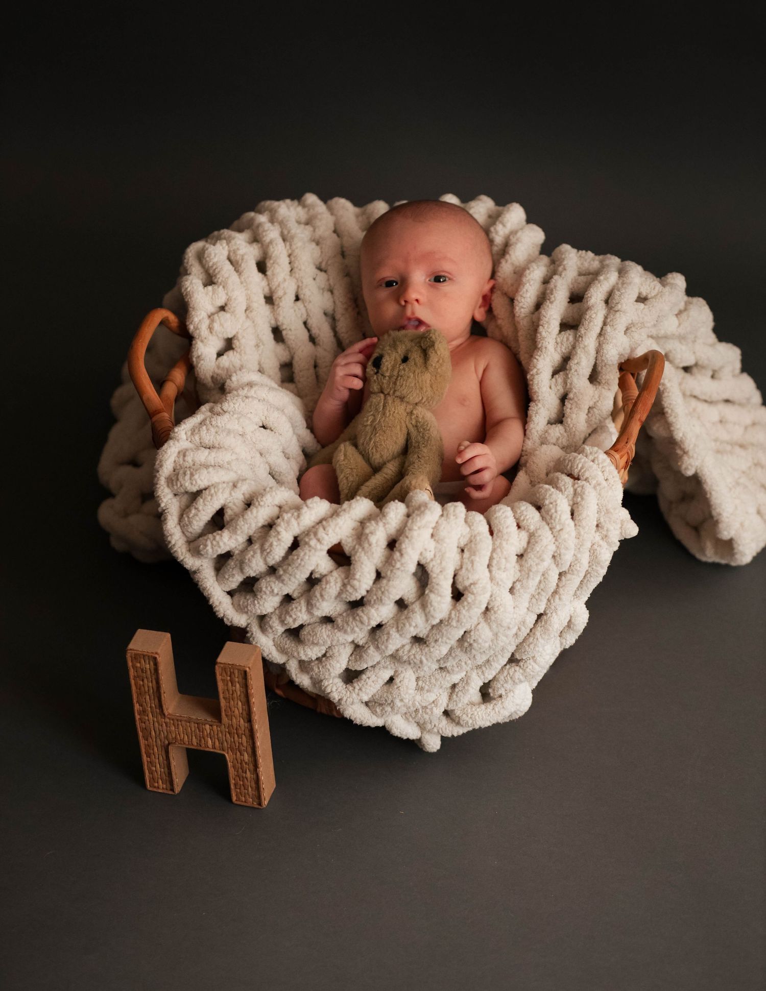 Newborn baby lying in chunky knit blanket with wooden letter H prop on dark background.
