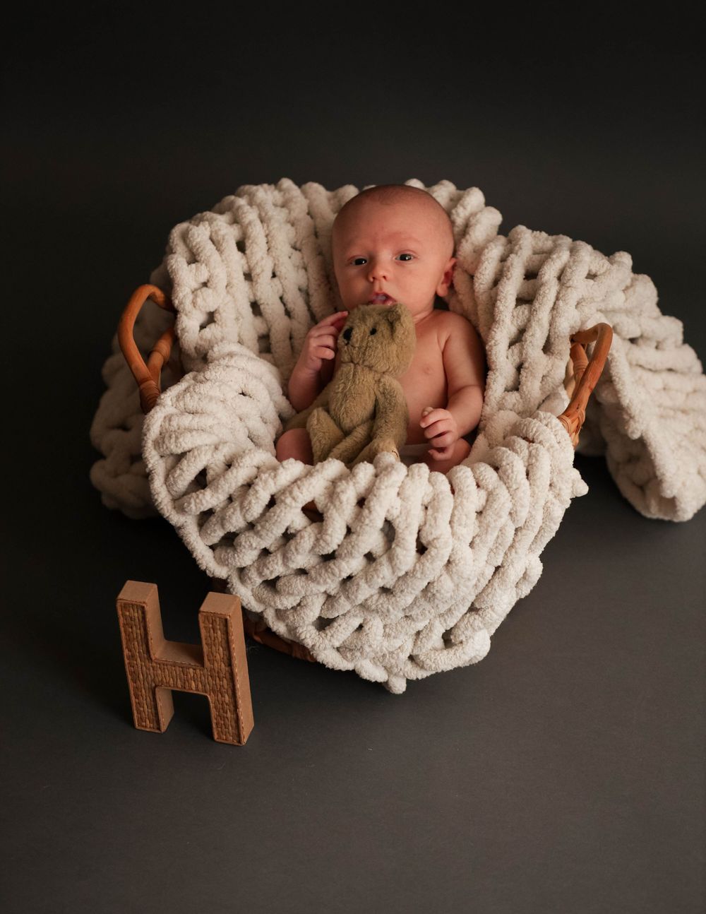 Newborn baby lying in chunky knit blanket with wooden letter H prop on dark background.