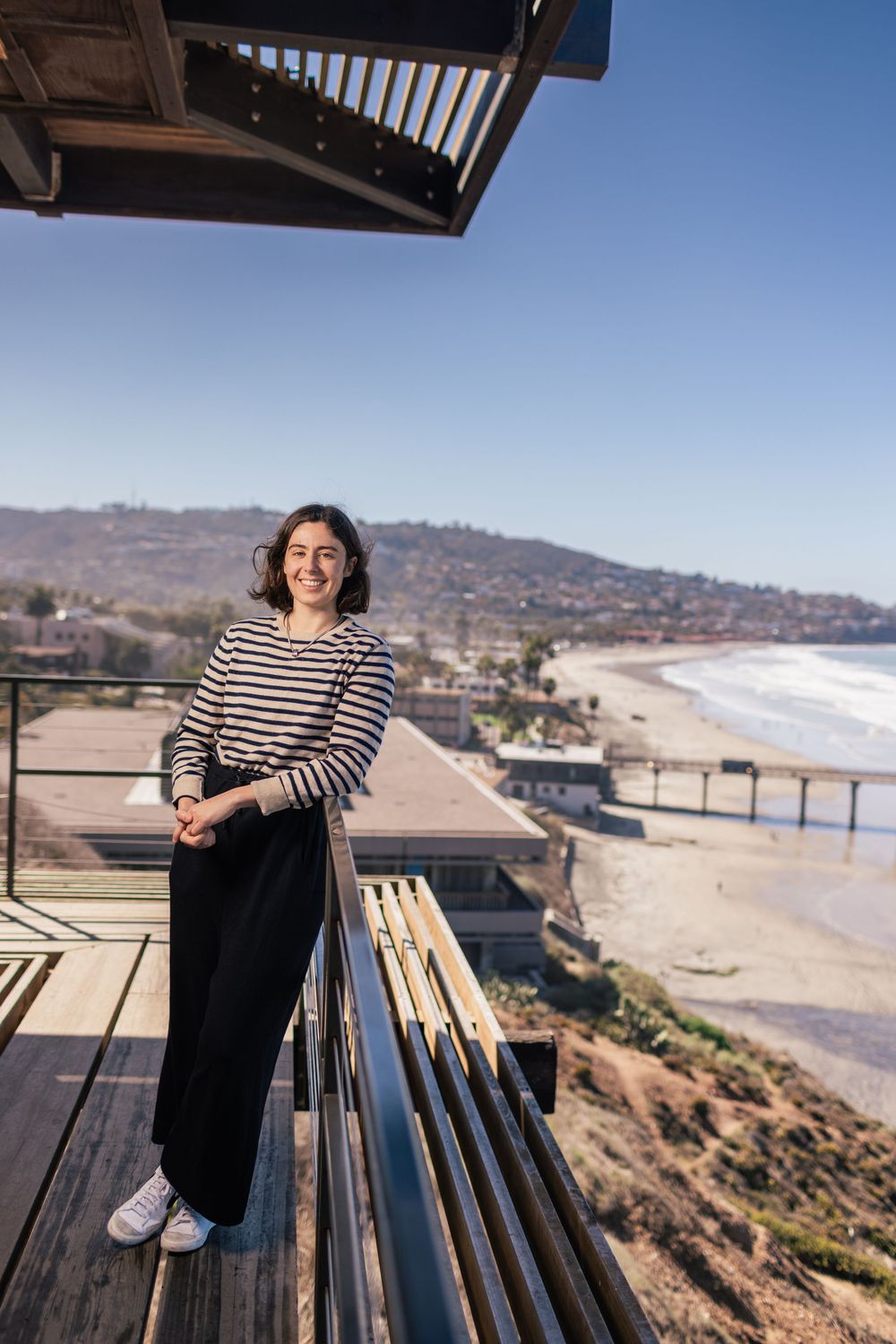 A person in a striped sweater stands on a wooden deck overlooking a scenic coastal beach on a sunny day.