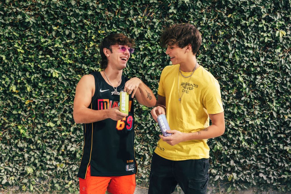 Two people in black and yellow outfits share a laugh while standing against a green hedge wall backdrop.