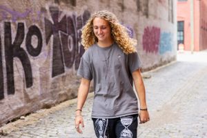 Person with curly blonde hair wearing grey t-shirt poses against urban graffiti wall on cobblestone street.