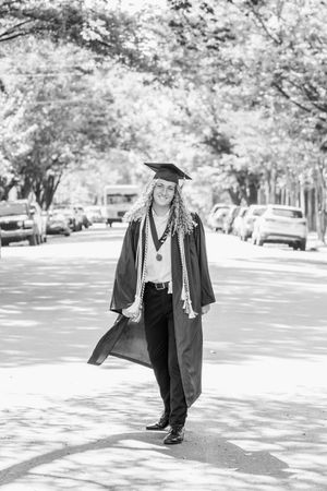 Graduate in cap and gown walking through tree-lined campus path in black and white.