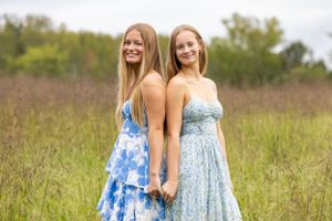 Two people in flowing summer dresses pose back-to-back in a sunny meadow.