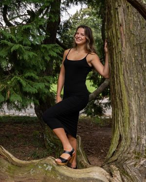 Person in black outfit poses playfully between large tree trunks in forest setting.