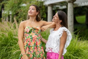Two people in floral and white dresses pose together in garden setting with pergola.