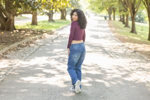Person strolls down sunny tree-lined path wearing casual denim and purple top.