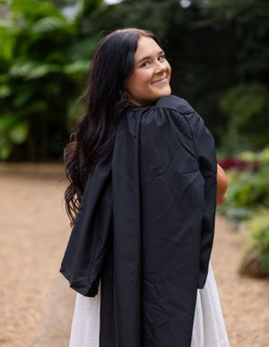 A person wearing graduation regalia smiles while posing in an outdoor garden setting.