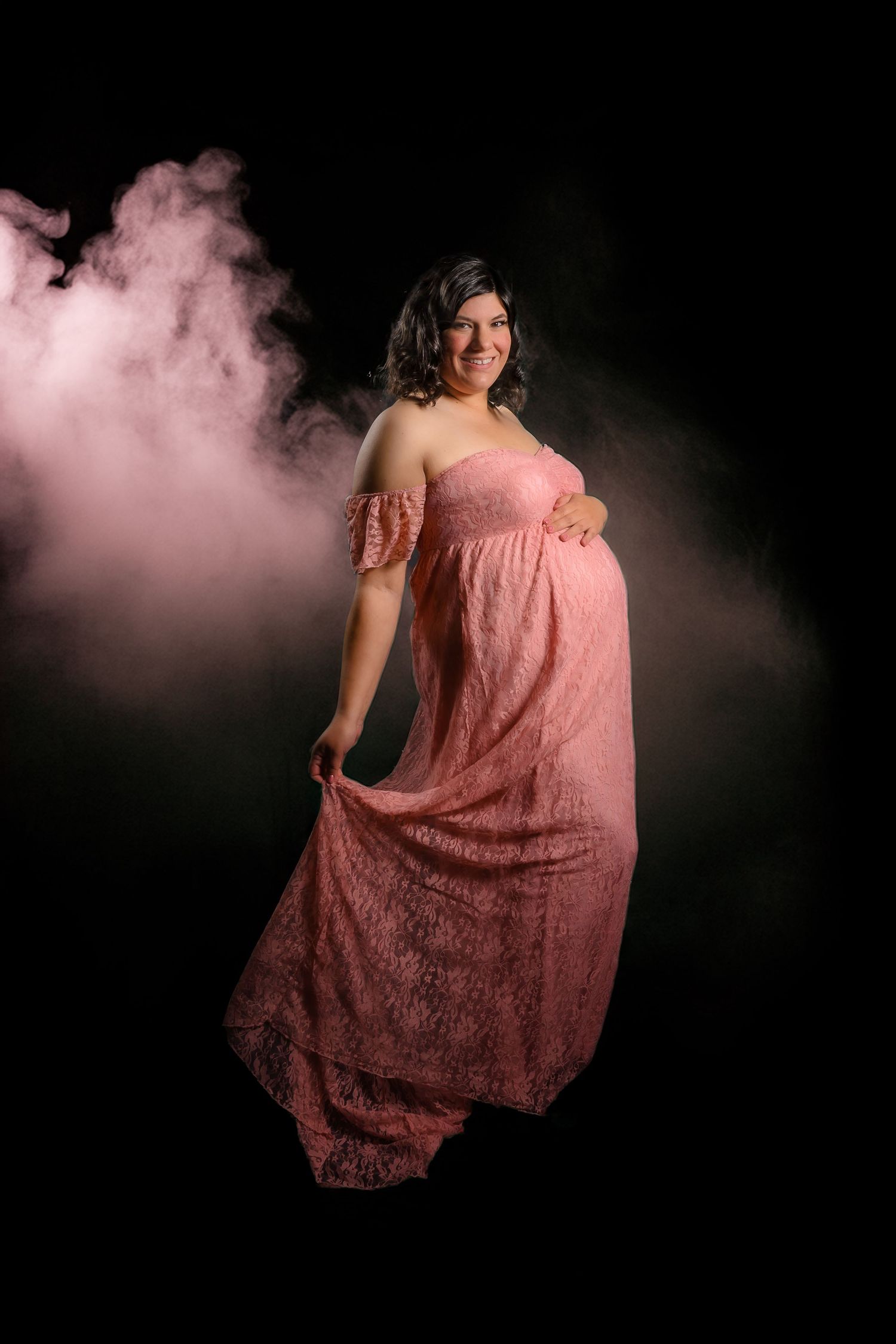 Smiling pregnant woman in pink lace maternity gown posed against dramatic smoky black background.