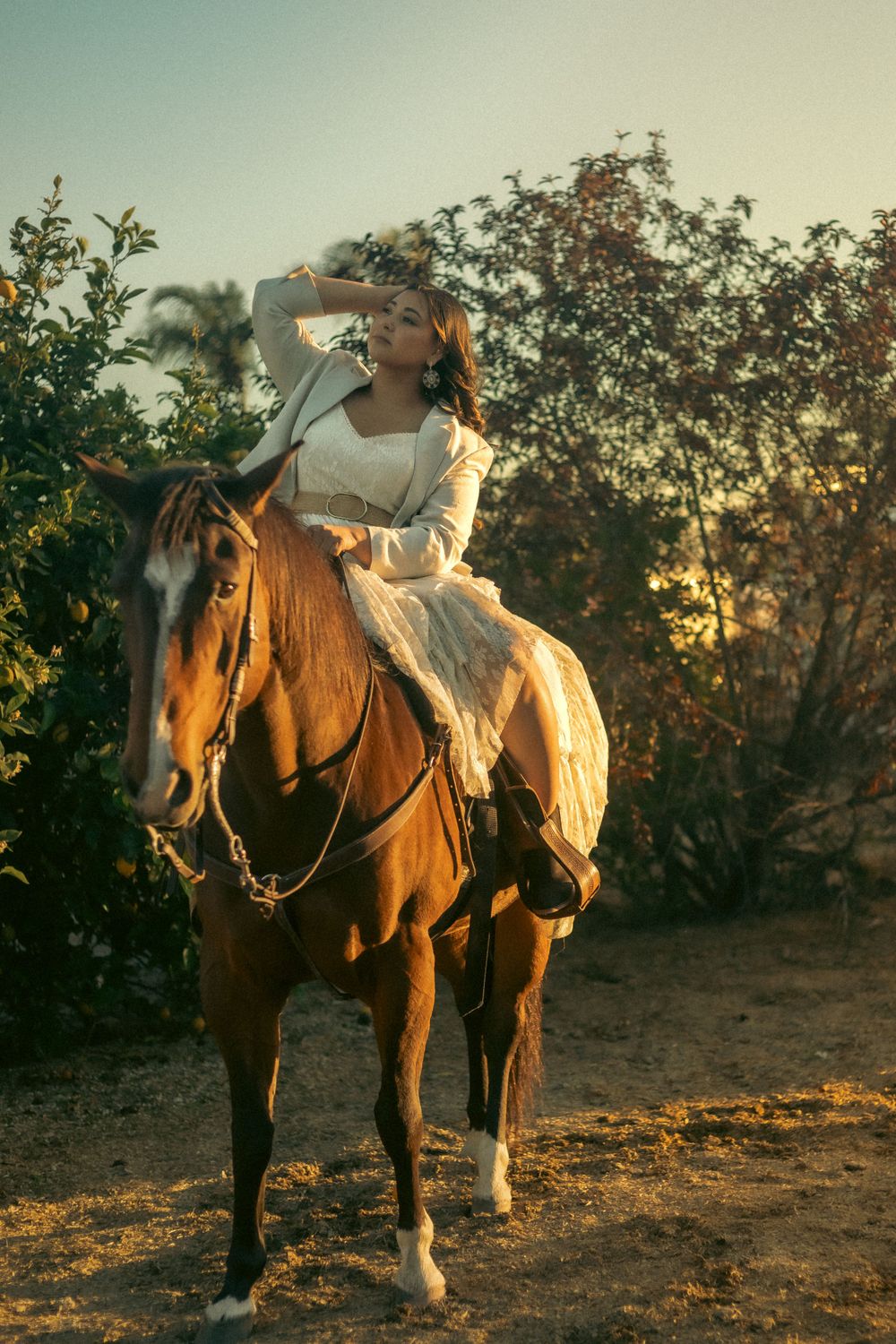 A rider in a flowing white dress sits atop a chestnut horse in golden sunset light for graduation photoshoot