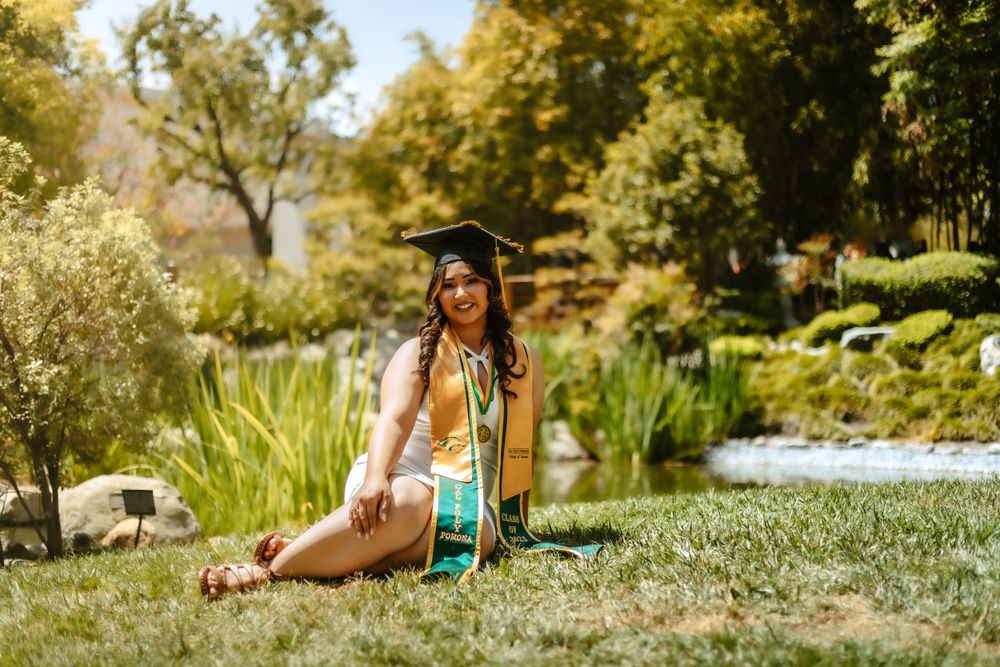 A graduate in cap and gown sits on grass field near Cal Poly pond in a sunny garden setting with lush trees and plants