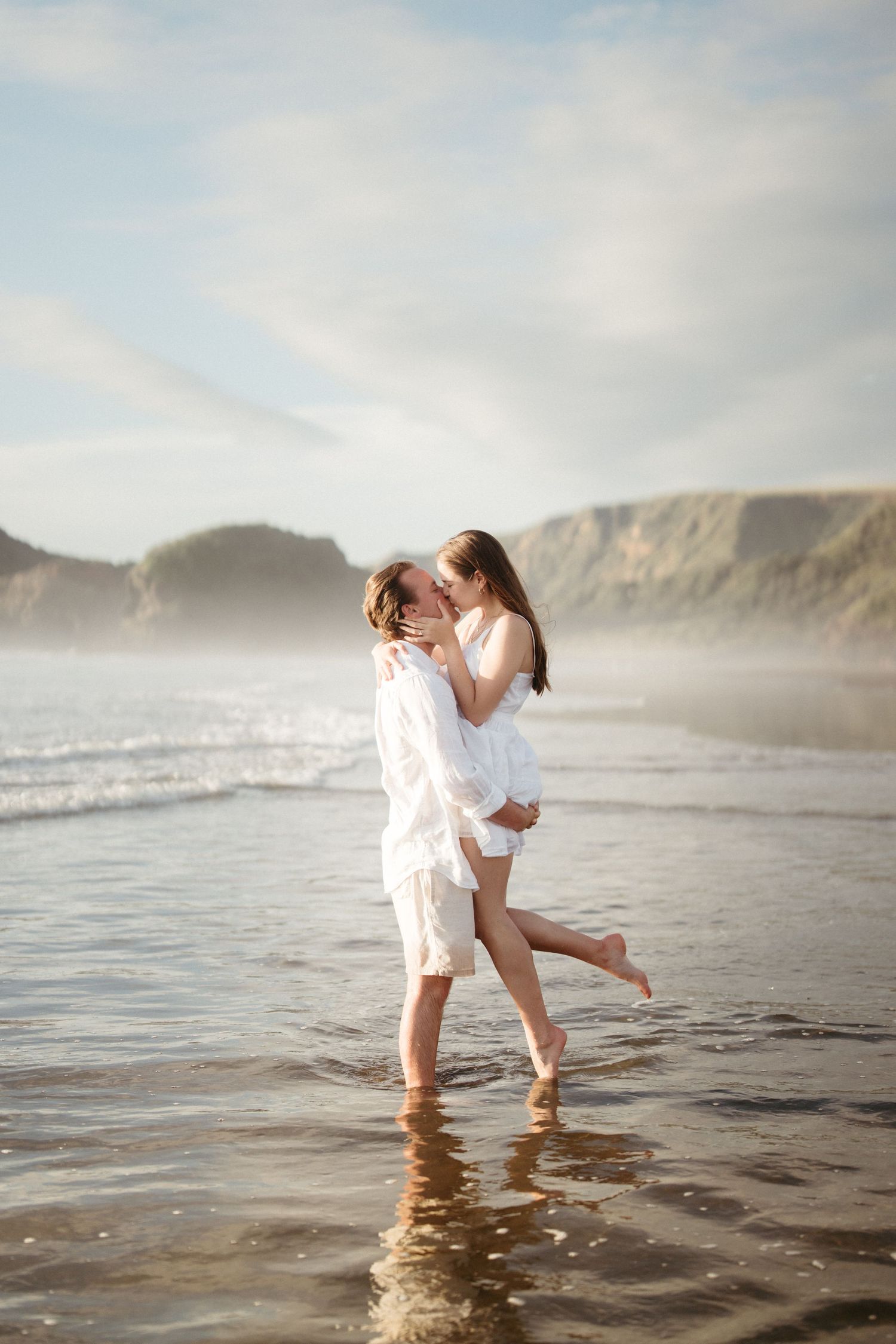 Couple embracing on misty beach with dramatic coastal cliffs in background on Bethells beach, Auckland.