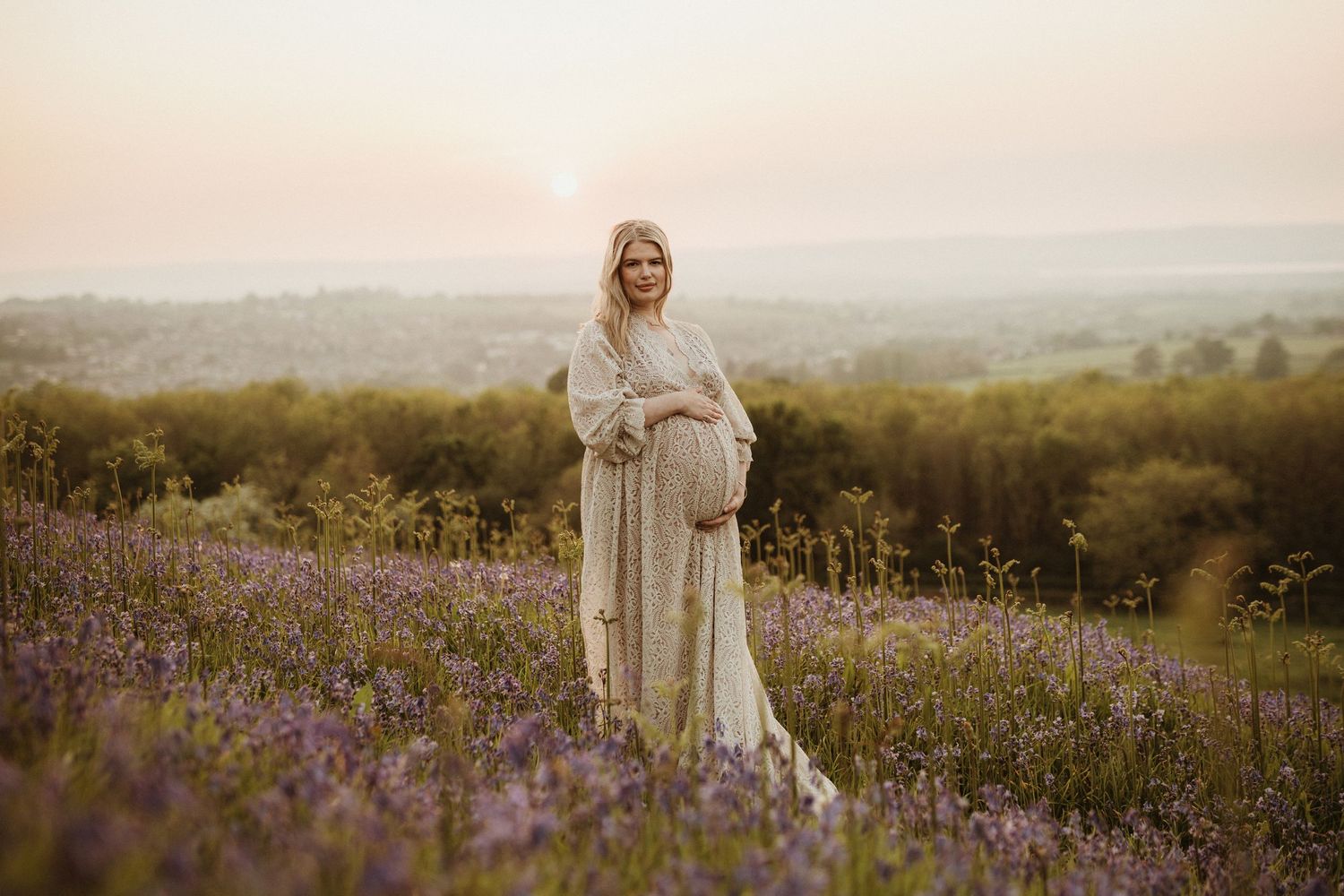 Ethereal maternity portrait in a flowing white dress against a sunset landscape.