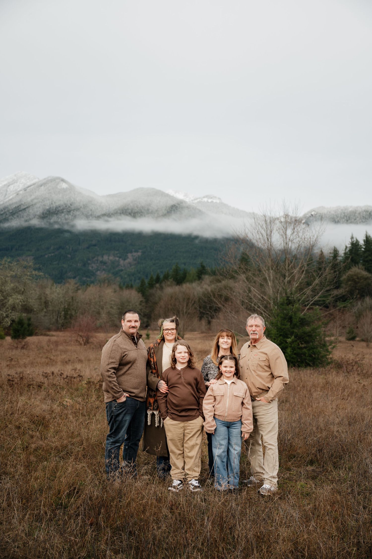 Couple posing in tall grass field with golden autumn trees and mountains in background.