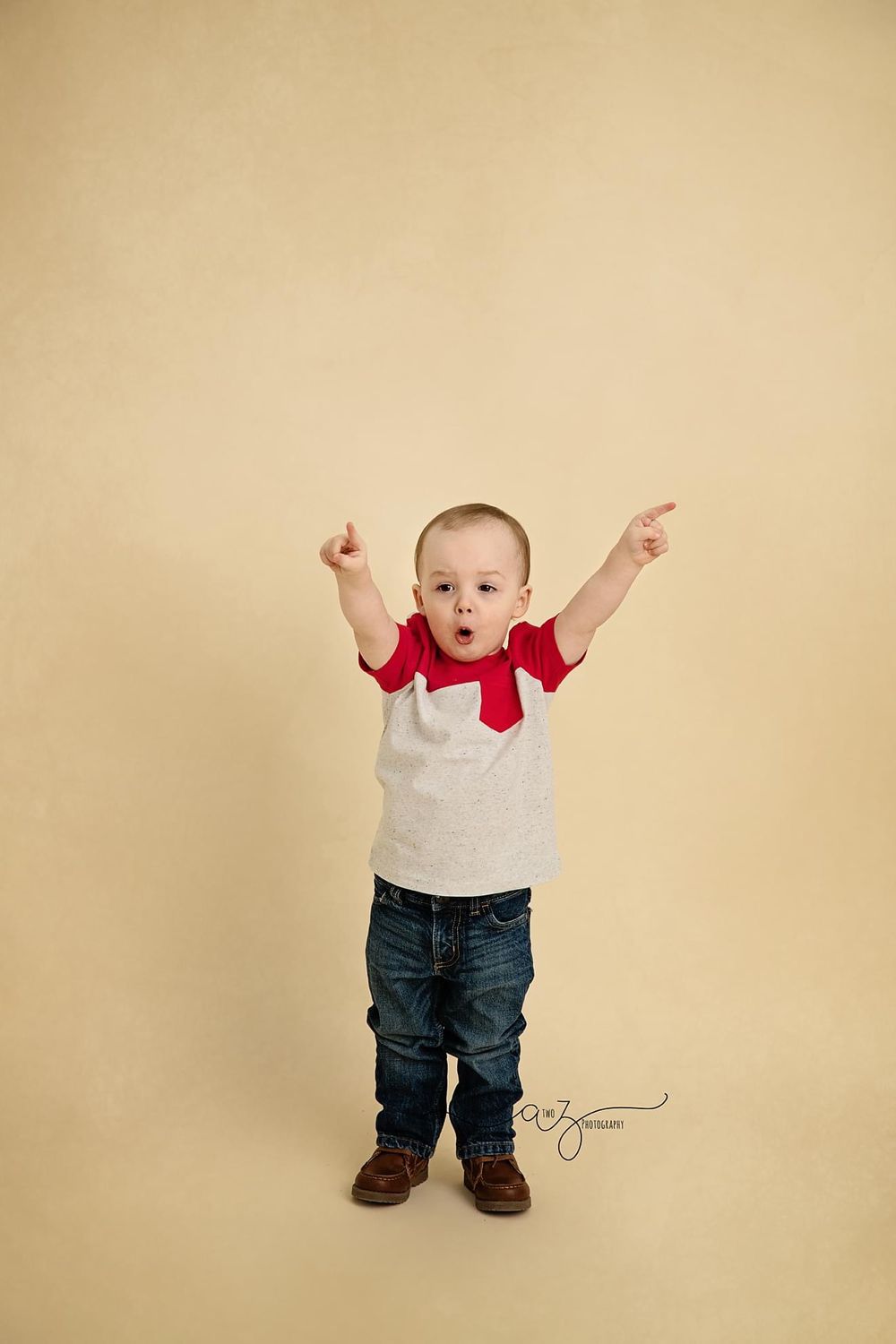Enthusiastic toddler in white shirt and red scarf raising arms against beige background.