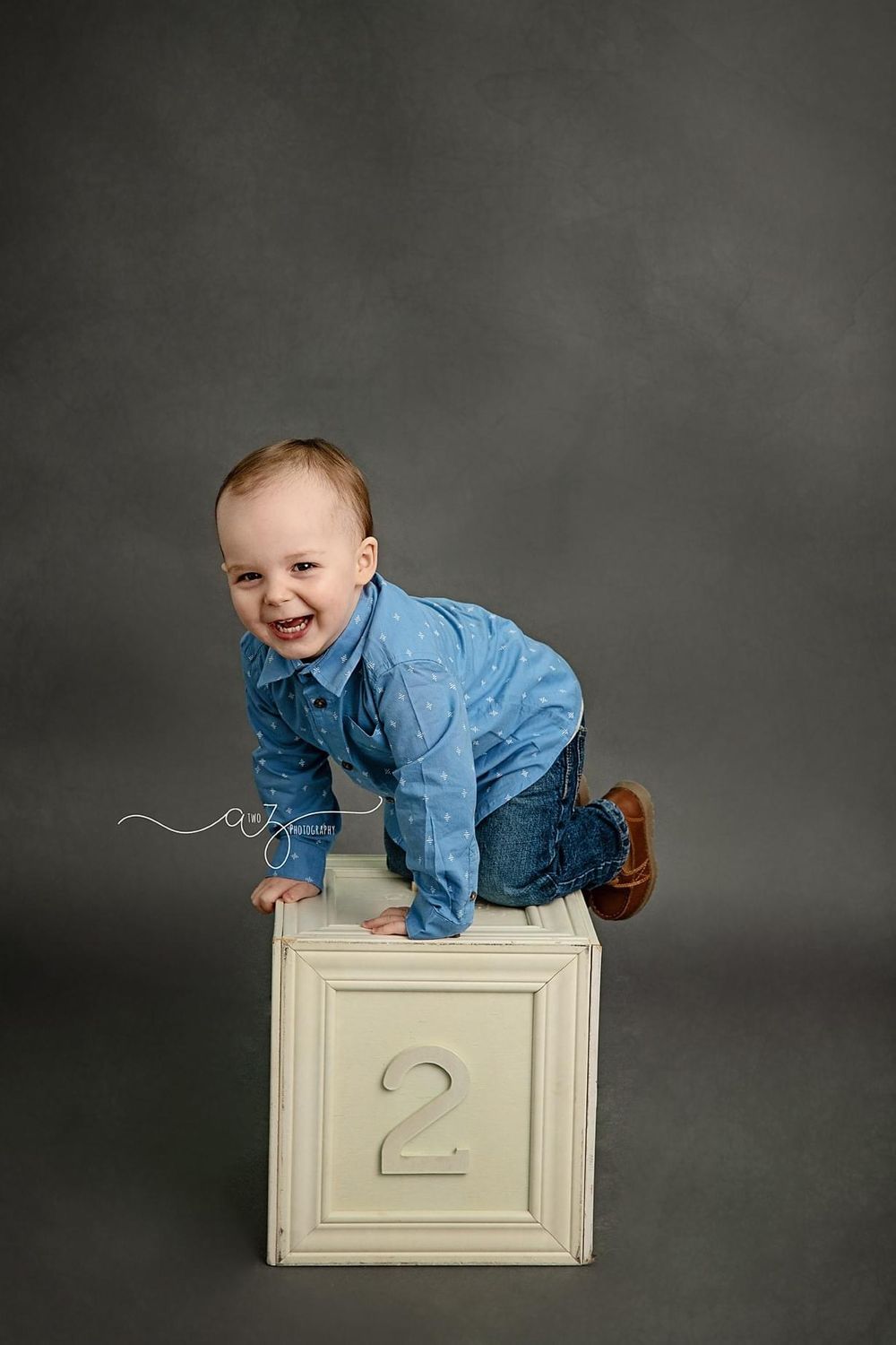 A smiling child in blue denim climbs on a white numbered cube against a grey backdrop.