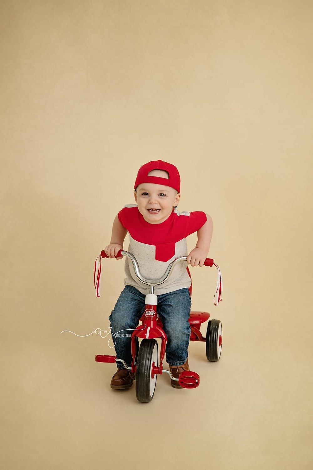A toddler wearing red and grey rides a red tricycle against a beige background.